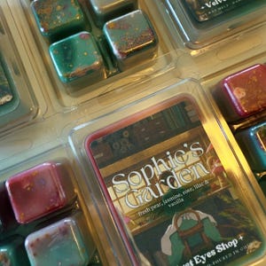 K&ouml;nnte beinhalten: Wachs-Schmelzw&uuml;rfel in einer transparenten Kunststoffverpackung. Die W&uuml;rfel sind in verschiedenen Farben gehalten, darunter Rot, Gr&uuml;n und Braun, mit gesprenkelten Akzenten. Auf dem Etikett steht "Sophie's Garden" mit Duftnoten und "Velvet Eyes Shop".