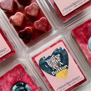 May include: Six heart-shaped wax melts in various shades of red and pink with glitter. The wax melts are in clear plastic containers. The text "You're Cute As A Button" is on a pink label with a cartoon image of a girl wearing a yellow jacket. The text "Velvet Eyes Shop" is on the label with the text "Soy Wax Melt - Hand-Poured in Ohio".