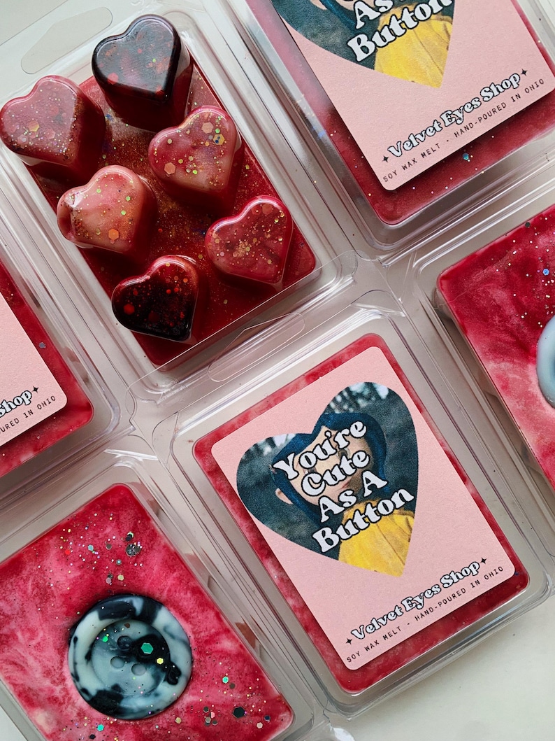 May include: Six heart-shaped wax melts in various shades of red and pink with glitter. The wax melts are in clear plastic containers. The text "You're Cute As A Button" is on a pink label with a cartoon image of a girl wearing a yellow jacket. The text "Velvet Eyes Shop" is on the label with the text "Soy Wax Melt - Hand-Poured in Ohio".