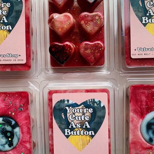May include: Six heart-shaped soy wax melts in a clear plastic package. The melts are red, pink, and white with glitter. The package has a label that says "You're Cute As A Button" and "Velvet Eyes Shop".