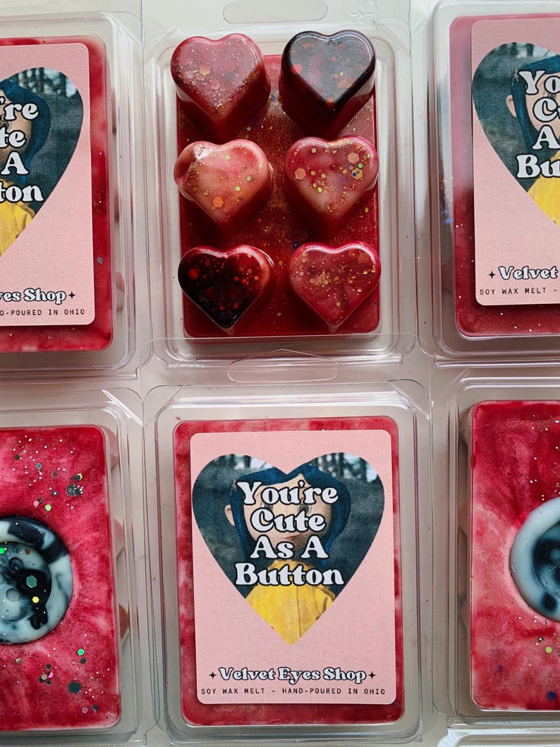 May include: Six heart-shaped soy wax melts in a clear plastic package. The melts are red, pink, and white with glitter. The package has a label that says "You're Cute As A Button" and "Velvet Eyes Shop".