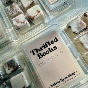 May include: Soy wax melt cubes in clear plastic packaging. The cubes are off-white with brown and red accents. The label reads "Thrifted Books" with scent notes. The packaging also includes the shop name and location.