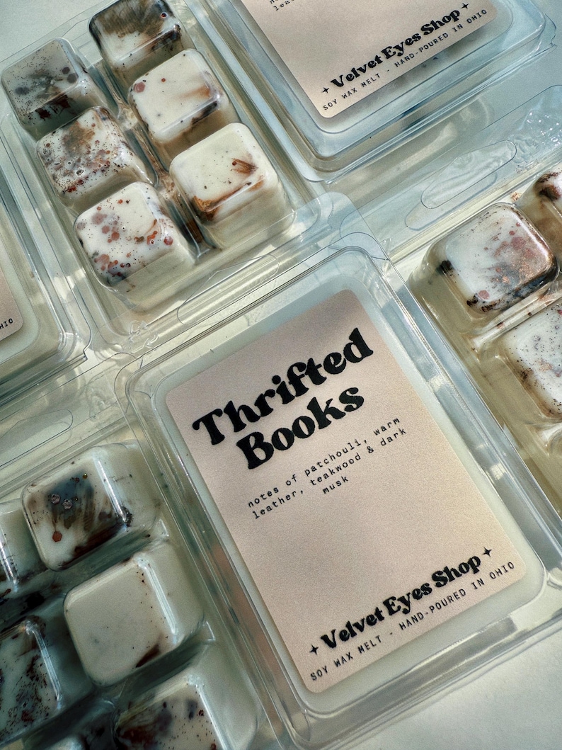May include: Soy wax melt cubes in clear plastic packaging. The cubes are off-white with brown and red accents. The label reads "Thrifted Books" with scent notes. The packaging also includes the shop name and location.