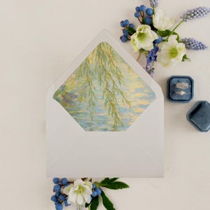 May include: A white envelope with a watercolor painting of a willow tree over water. The envelope is surrounded by white flowers, blue berries, and two rings in a blue velvet box. Wedding stationery.