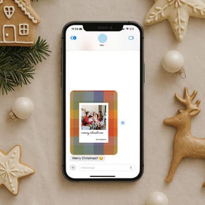May include: A smartphone displays a Christmas message with a family photo, surrounded by festive decorations. Gingerbread house, star-shaped cookies, ornaments, and a reindeer figurine add to the holiday theme. The message reads "Merry Christmas!!"