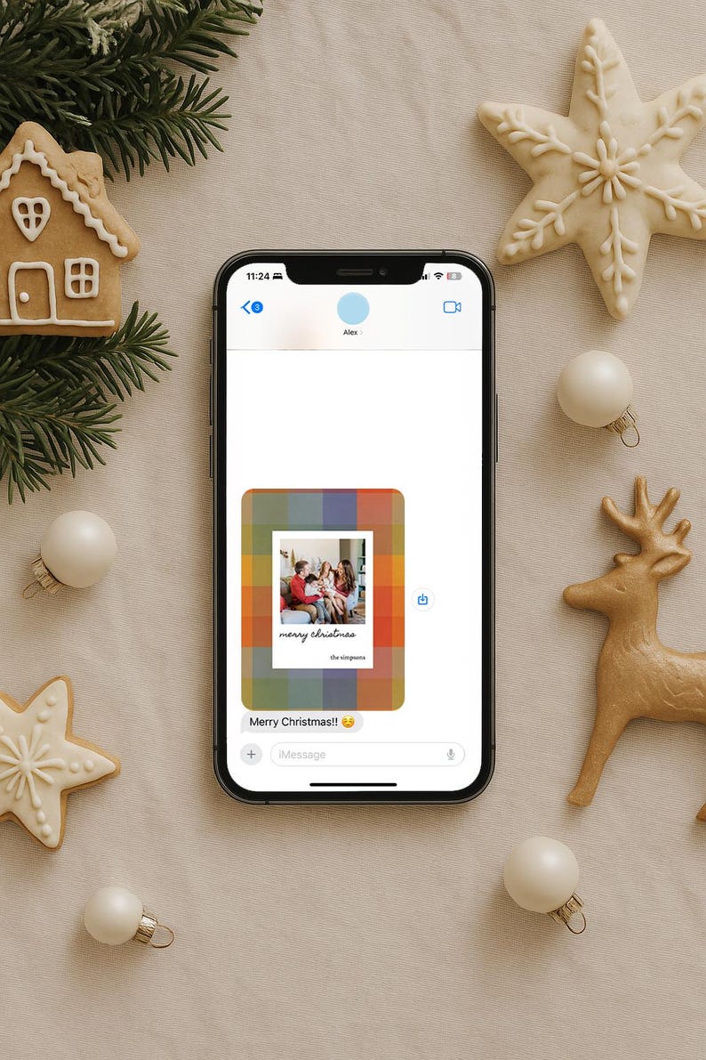 May include: A smartphone displays a Christmas message with a family photo, surrounded by festive decorations. Gingerbread house, star-shaped cookies, ornaments, and a reindeer figurine add to the holiday theme. The message reads "Merry Christmas!!"
