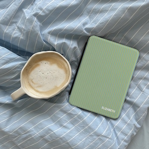 Könnte beinhalten: Eine Tasse Kaffee in einer weißen Tasse und eine grün-blau gestreifte E-Reader-Hülle mit dem Namen "ELIZABETH" darauf, auf einem blau-weiß gestreiften Bettlaken.