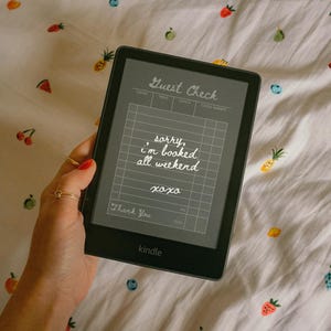 Op de afbeelding: Een zwarte Kindle e-reader met een aangepast "Guest Check" ontwerp met de tekst "sorry, I'm booked all weekend xoxo". De e-reader ligt op een witte stof met geborduurde fruitpatronen.