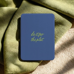 May include: A rectangular blue card with vertical stripes and rounded corners. The card features the phrase "do it for the plot" in a handwritten-style green font. The card is resting on a green and tan fabric.