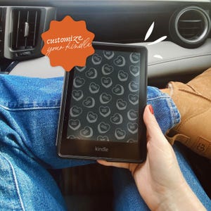 May include: A black Kindle e-reader with a custom screen saver featuring a pattern of small white hearts on a black background. The text "customize your kindle" is written on a bright orange starburst sticker.