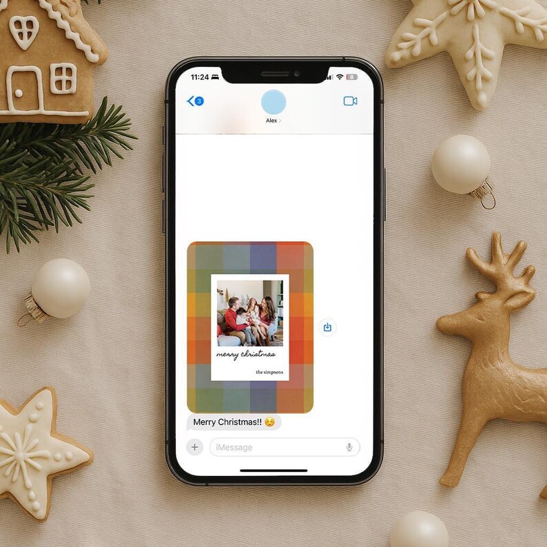 May include: A smartphone displays a Christmas message with a family photo. The message is framed with a colorful plaid pattern. The text reads "Merry Christmas!!" and "the simpsons". The phone is surrounded by holiday decorations.
