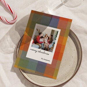 May include: Christmas card with a family photo inside a white frame, set against a plaid pattern in orange, green, and blue. The card reads "merry christmas" and "the simpsons". A candy cane rests on the side.