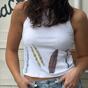 May include: White ribbed vest top with colourful, stitched leaf-like designs. The top is paired with light-wash denim shorts. The shorts have a classic five-pocket design and a button and zip closure.