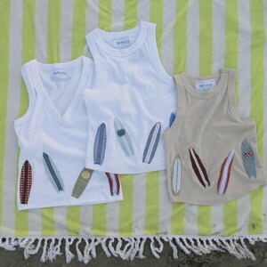 May include: Three sleeveless tank tops featuring various surfboard-shaped fabric appliqués. Two tops are white with a ribbed texture, one with a V-neck and one with a crew neck. The third top is a solid beige color with a crew neck and surfboard patches.