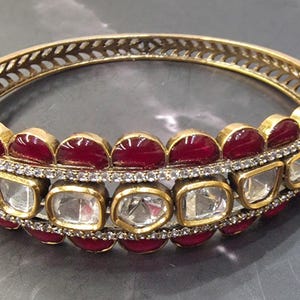 May include: A gold-toned hinged bangle bracelet featuring a central row of clear, square-cut stones bordered by two rows of deep red, semi-circular cabochon stones. The metal frame displays a delicate, openwork leaf-like pattern along the inner edge.