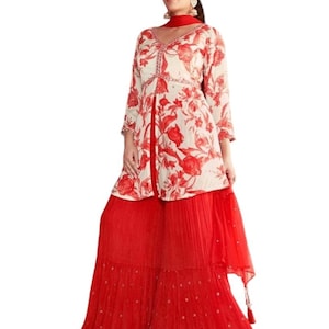 May include: A two-piece outfit featuring a cream-colored top with a red floral pattern and a matching red palazzo pant. The top has a V-neck and three-quarter sleeves. The palazzo pants are wide-legged and flowing, with a decorative trim.