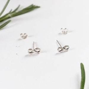 Op de afbeelding: Een paar zilverkleurige infinity-symbool oorknopjes met twee oorbelruggen. De oorbellen liggen op een witte ondergrond met een takje groene bladeren op de achtergrond.