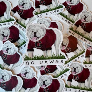 May include: A collection of stickers featuring a white bulldog wearing a maroon jersey with a black collar and the letter "G". The stickers have the text "GO DAWGS" and are surrounded by green grass.