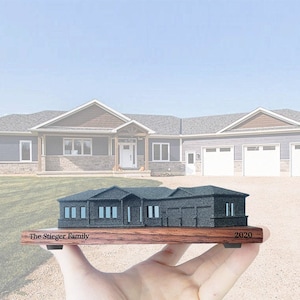 May include: A miniature replica of a house on a wooden base with the inscription "The Steiger Family 2020". The replica is painted grey and has a white front door. The miniature house is being held in a person's hand.