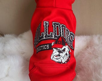 Può includere: Una felpa con cappuccio rossa brillante per animali domestici con la grafica di un bulldog che indossa un berretto rosso e nero. Il testo "BULLDOGS" è stampato in grandi lettere bianche con un bordo nero sopra la parola "ATHLETICS".