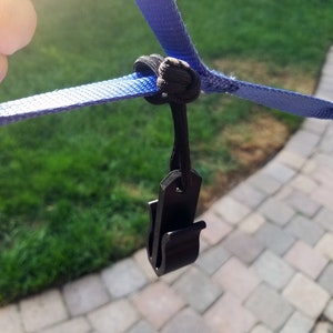 May include: A close-up of a black plastic clip attached to a blue strap. The strap is knotted with a black cord. The background shows green grass and a paved surface.
