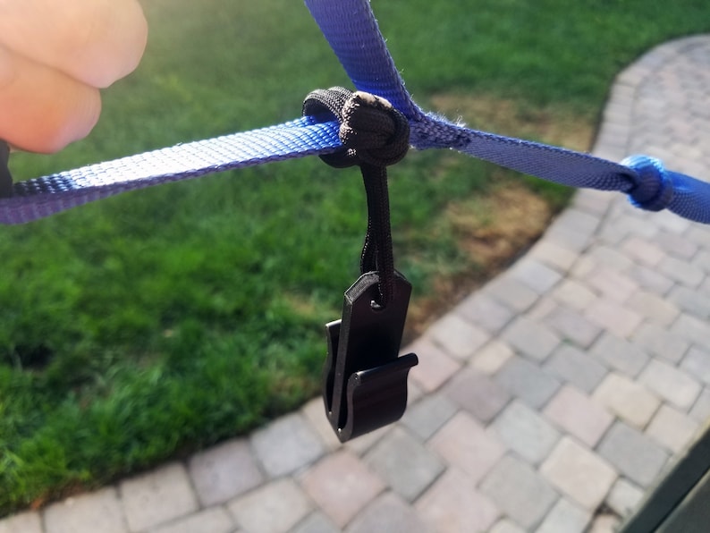 May include: A close-up of a black plastic clip attached to a blue strap. The strap is knotted with a black cord. The background shows green grass and a paved surface.