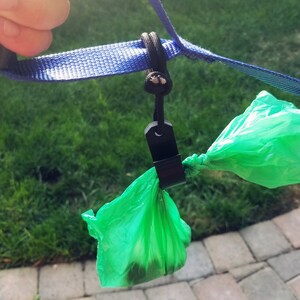 May include: A blue dog leash with a black clip holding two green waste bags. The leash is held by a hand, with a grassy background and a brick walkway visible.