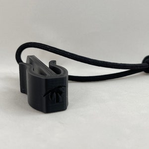May include: A black 3D-printed cord lock with a palm tree design. The cord lock is attached to a black elastic cord. The cord lock has two curved arms and a slot for the cord.
