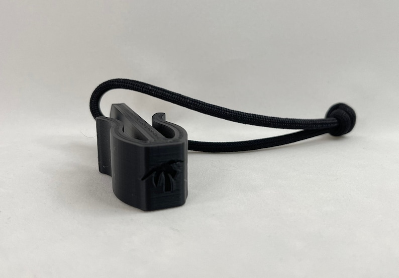 May include: A black 3D-printed cord lock with a palm tree design. The cord lock is attached to a black elastic cord. The cord lock has two curved arms and a slot for the cord.