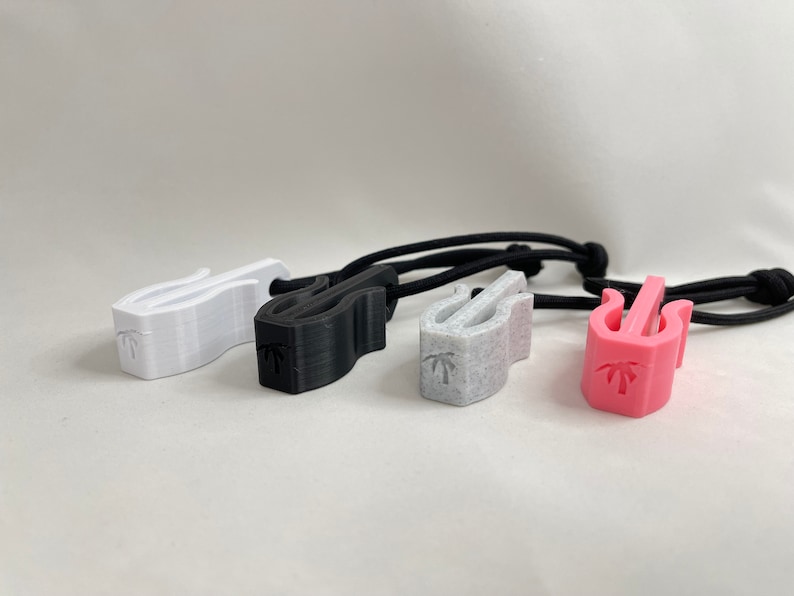 May include: Four 3D-printed cord keepers in white, black, speckled gray, and pink. Each has a curved design with a palm tree logo and is attached to a black elastic cord. These are designed to manage and organize cords.