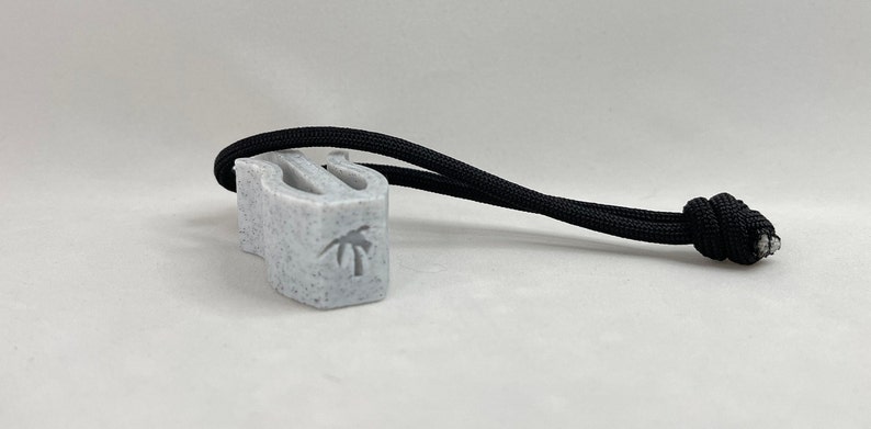 May include: A speckled gray whistle with a black cord attached. The whistle has a palm tree design etched on the side. The black cord is knotted at the end.