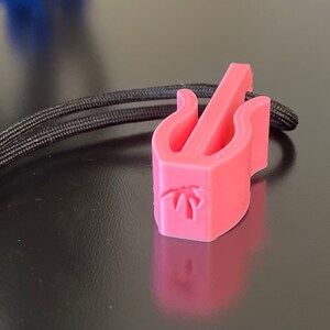 May include: A pink 3D-printed guitar pick holder with a black cord. The holder has a stylized design with a palm tree graphic. The cord is knotted at one end.