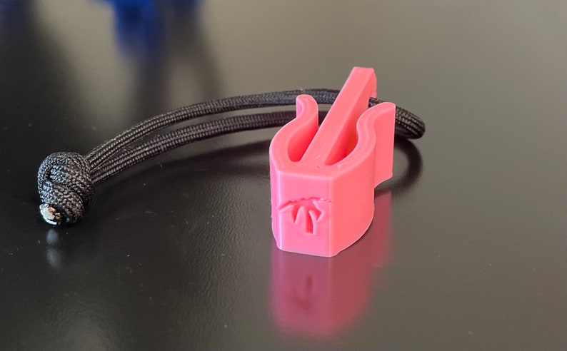 May include: A pink 3D-printed guitar pick holder with a black cord. The holder has a stylized design with a palm tree graphic. The cord is knotted at one end.