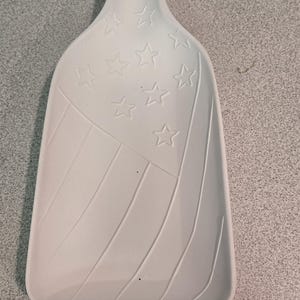 May include: A white ceramic spoon rest shaped like a bottle with a flat base and a narrow neck. The surface features an embossed design of stars in the upper section and diagonal stripes in the lower section, mimicking a flag pattern in relief.
