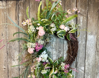 May include: A pink and green floral wreath with a brown twig base. The wreath is decorated with pink roses, white flowers, and green leaves.