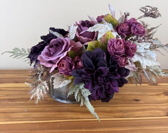Könnte beinhalten: Ein Blumenarrangement in einer Glasschale mit dunkelvioletten Dahlien, hellvioletten Rosen und kleineren tief magentafarbenen Blüten. Das Gesteck enthält silberfarbenes farnartiges Laub, weiß gespitzte Blätter und kleine weiße Beeren.