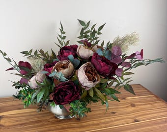 Könnte beinhalten: Ein Blumenarrangement mit tiefburgunderfarbenen und gedeckten malvenfarbenen Pfingstrosen in einer silberfarbenen Metallschale. Der Strauß enthält diverses grünes Blattwerk, feine Trockengräser und eukalyptusartige Blätter, die ein dichtes Arrangement bilden.