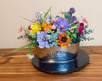 May include: A colorful artificial flower arrangement in a silver metal container with the words "Flowers & Garden" on the side. The arrangement includes purple, yellow, orange, pink, and green flowers.
