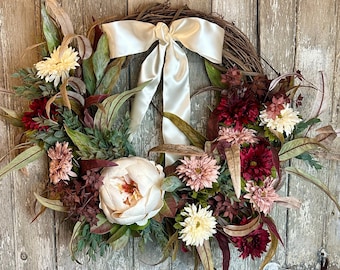 May include: A wreath with a white satin bow and a variety of faux flowers in shades of pink, white, and burgundy. The wreath is made of grapevine and is decorated with greenery.