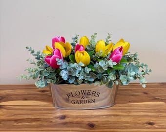 May include: A floral arrangement featuring vibrant pink and yellow tulips, accented by green foliage. The flowers are arranged in a wooden oval planter with the words "FLOWERS & GARDEN" printed on the front. The arrangement sits on a wooden surface.