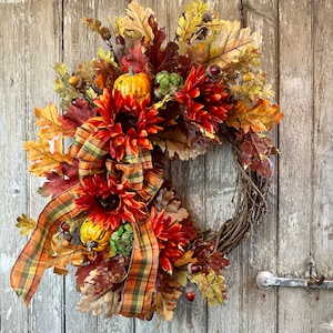 May include: A fall-themed wreath with orange and yellow gourds, red and orange flowers, and brown leaves. The wreath is made of twigs and has a plaid ribbon tied around it.