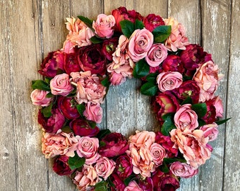 May include: A pink and burgundy floral wreath made of silk flowers. The wreath is made of roses, peonies, and other flowers. It is a beautiful and elegant wreath that would be perfect for a wedding or other special occasion.