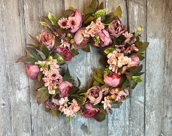 May include: A pink and green floral wreath with various types of flowers, including peonies, roses, and hydrangeas. The wreath is hanging on a wooden door.