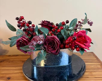 May include: A floral arrangement featuring deep red roses, berries, and greenery in a galvanized metal container. The container has the words "Flowers & Garden" printed on it. The arrangement sits on a dark gray, round base, and is displayed on a wooden surface.