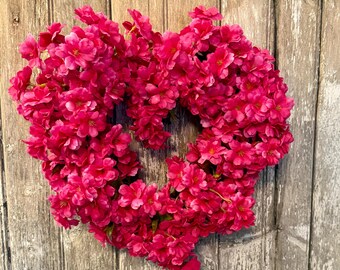 May include: A vibrant pink floral wreath, heart-shaped, composed of densely packed blossoms. The wreath is set against a weathered, gray wooden background, creating a striking contrast. The flowers are a bright, saturated pink.