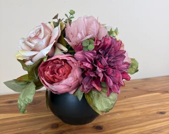 Könnte beinhalten: Ein Blumenarrangement aus künstlichen Blüten in Altrosa, tiefem Magenta und Burgunderrot, akzentuiert durch grüne Blätter und kleine Zweige, untergebracht in einer glatten, mattschwarzen, abgerundeten schalenförmigen Vase.