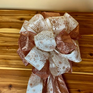 May include: A white and rose gold Christmas tree topper bow with a snowflake pattern. The bow is made of two different types of ribbon, one with a sheer texture and the other with a glitter finish.