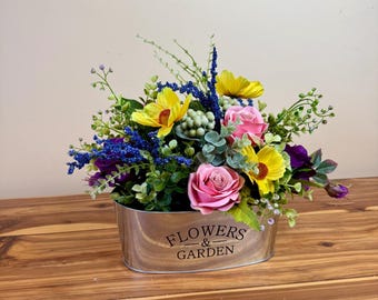 May include: An oval metal container filled with a vibrant arrangement of artificial flowers. The arrangement includes yellow sunflowers, pink roses, and blue and purple accents. The container has the words "FLOWERS & GARDEN" printed on the front.