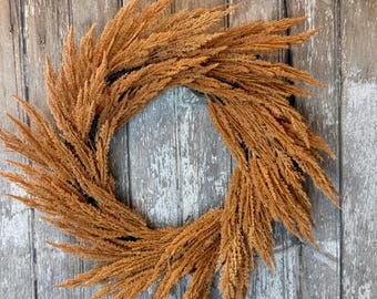 May include: A dried pampas grass wreath in a natural brown color. The wreath is hanging on a rustic wooden background.