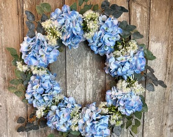 May include: A blue and white hydrangea wreath with eucalyptus leaves. The wreath is made of artificial flowers and is perfect for spring or summer decor.